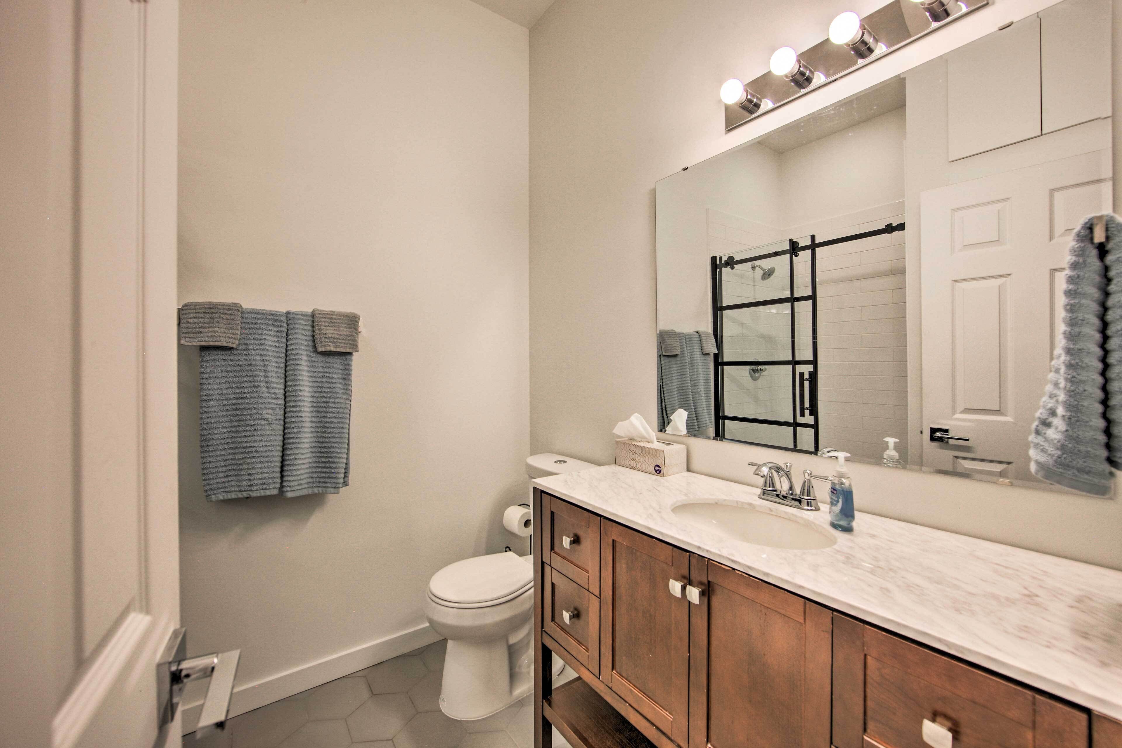 Un baño con inodoro blanco, un lavabo con encimera de mármol, armarios de madera y un espejo grande con luces arriba.