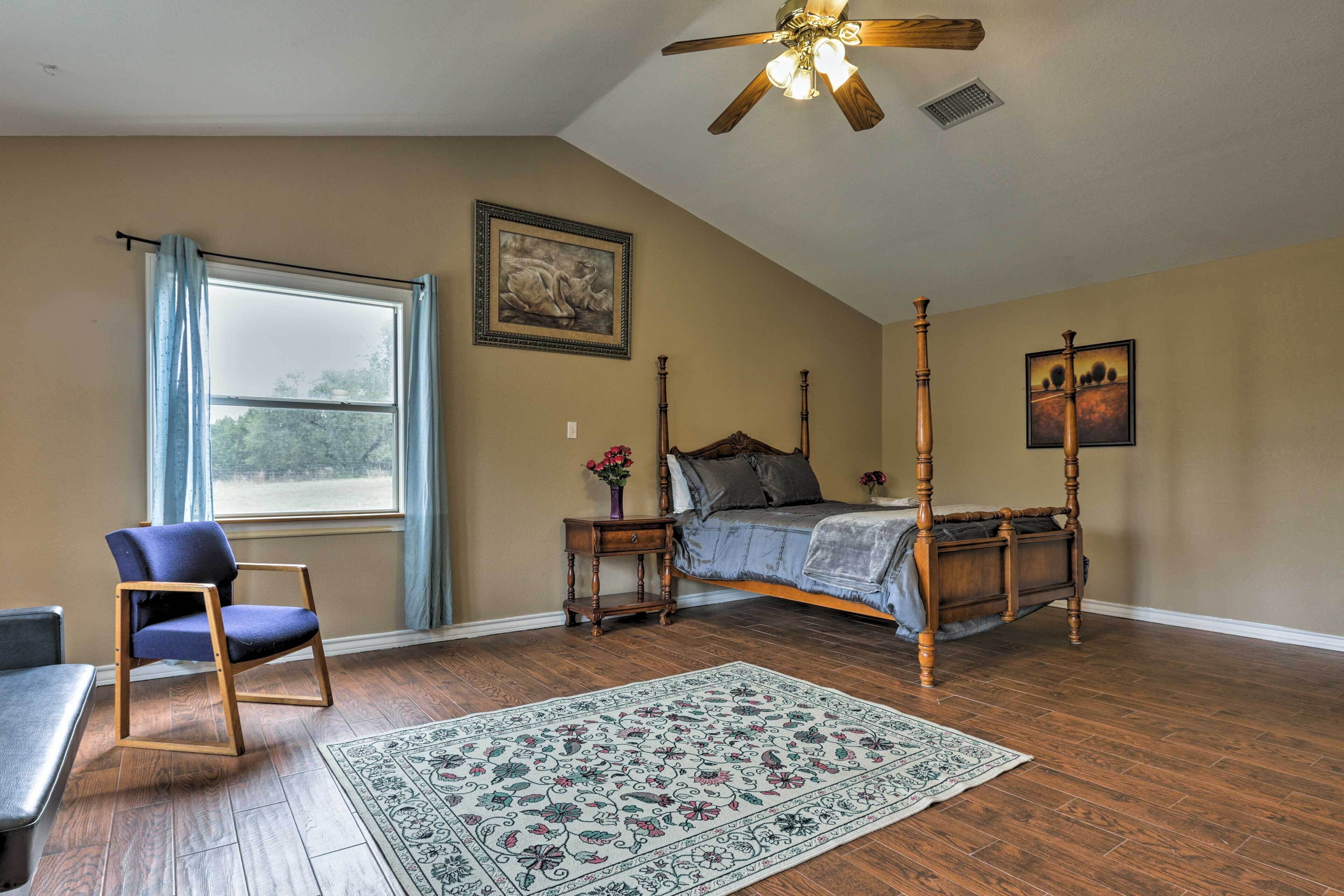 A bedroom with a four-poster bed, a wooden chair, a desk, a large window with curtains, and a ceiling fan.