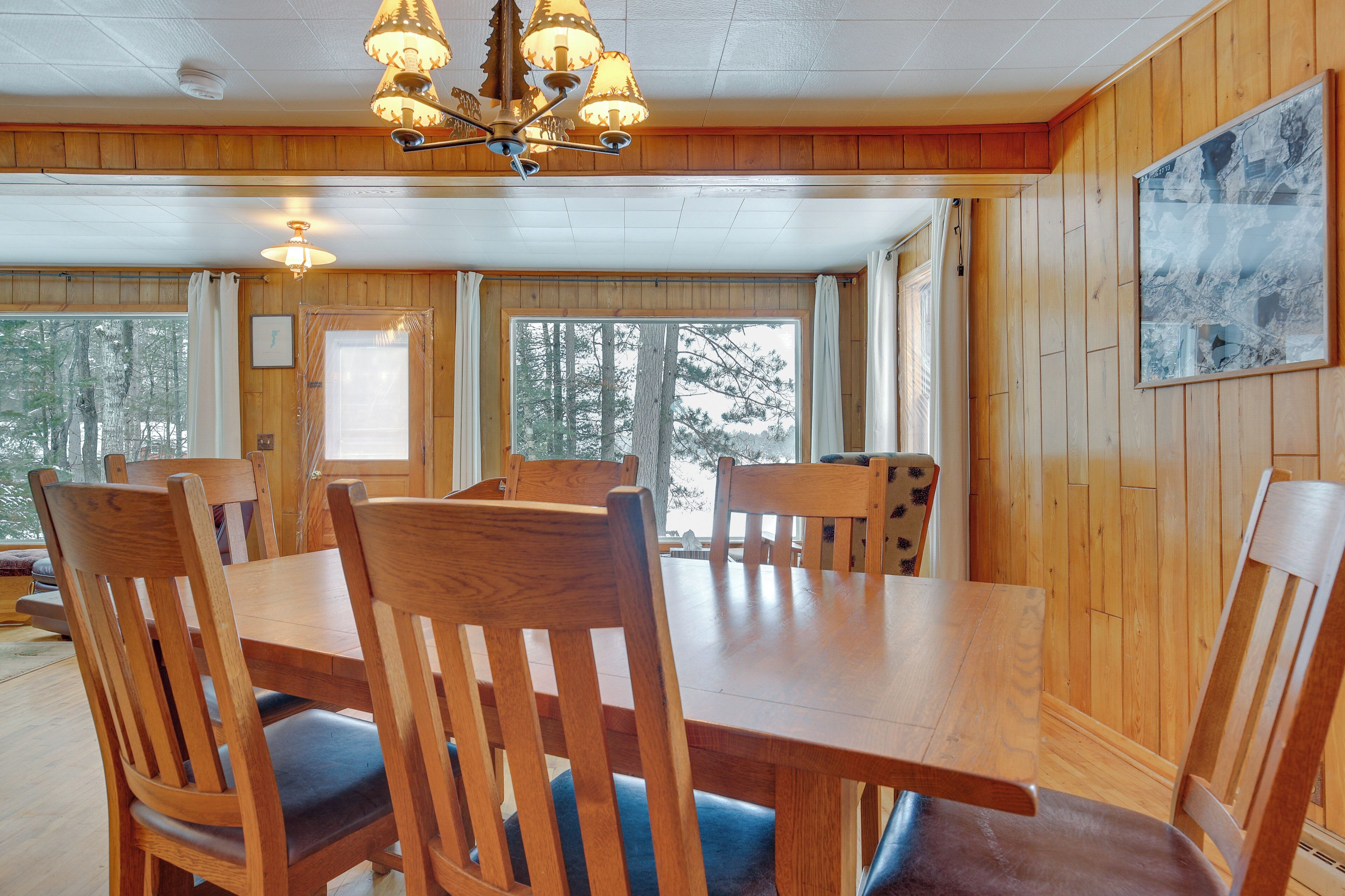 A wooden-paneled dining area with a large table and chairs, a chandelier, and a window with a view of trees.