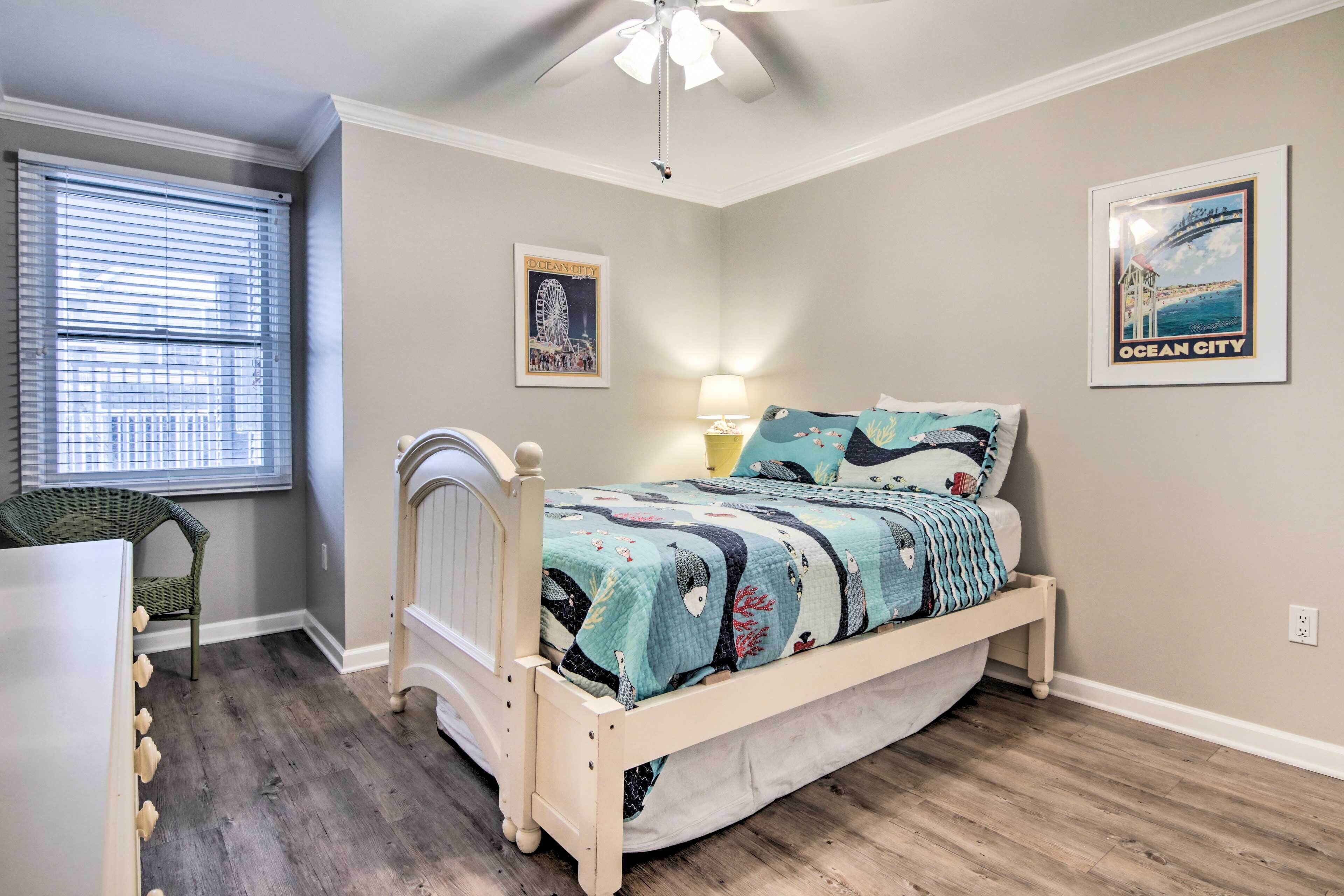 Une chambre à coucher comprenant un lit, une chaise, une fenêtre avec des stores, un ventilateur de plafond et deux tableaux encadrés accrochés au mur.
