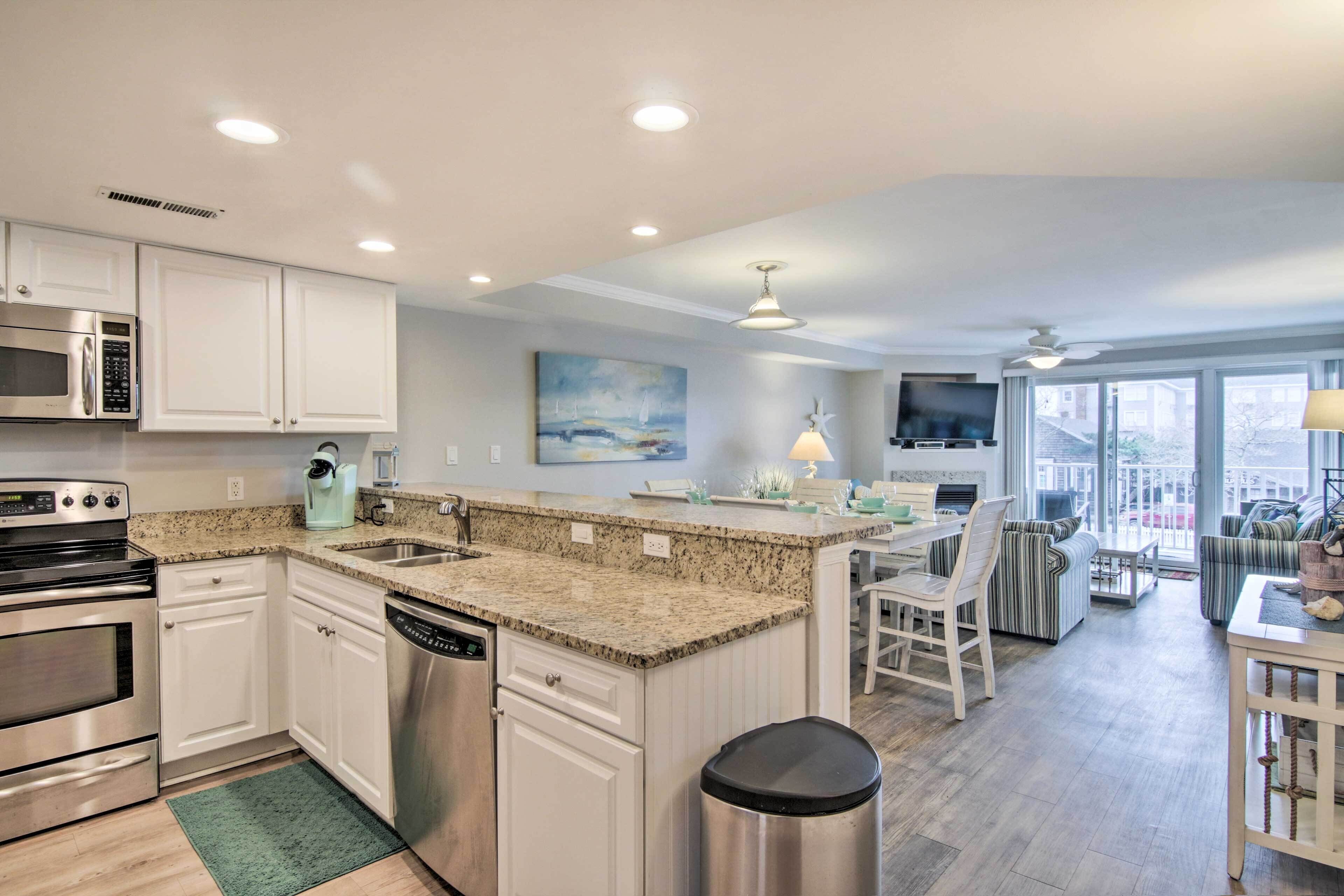 A modern kitchen with white cabinets, granite countertops, stainless steel appliances, and a dining area with striped chairs and a TV.