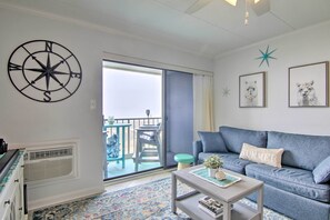A living room with a blue sofa, a coffee table, a large window, and a decorative compass wall art. - Condo Retreat w/ Balcony on Ocean City Beach! (Ocean City)