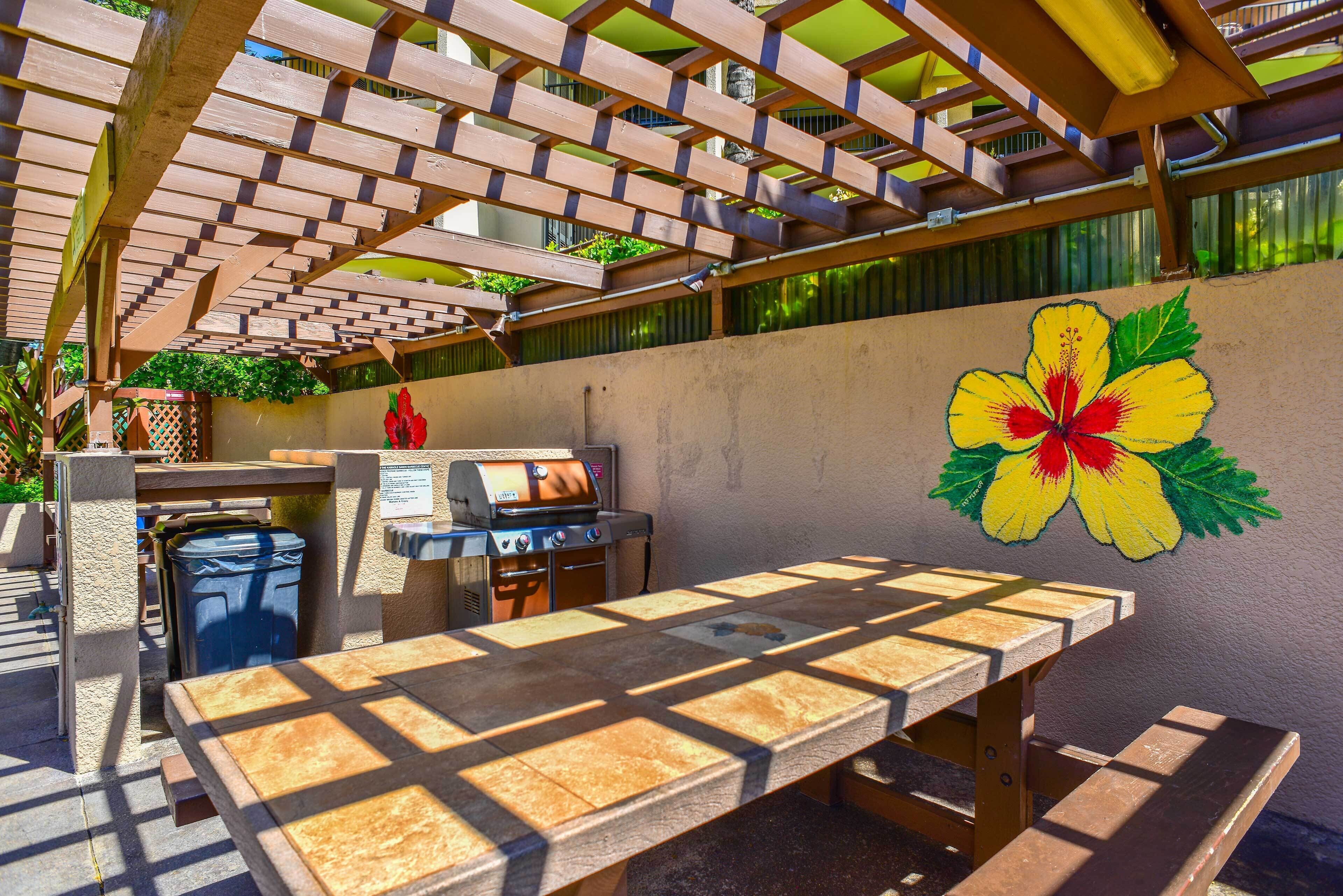 An outdoor patio with a wooden table and chairs, a barbecue grill, and a hibiscus flower mural on the wall.