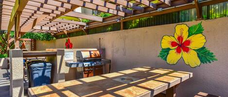 An outdoor patio with a wooden table and chairs, a barbecue grill, and a hibiscus flower mural on the wall.