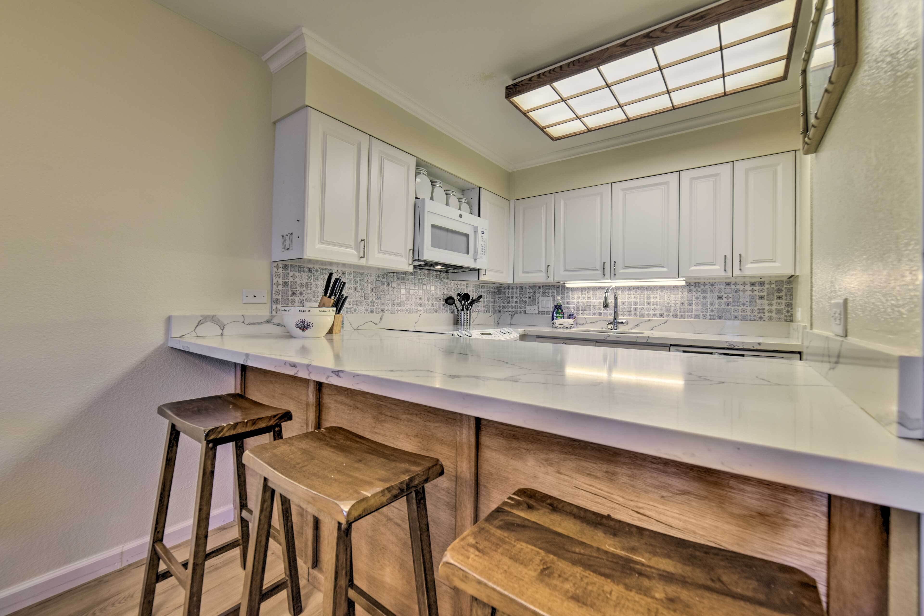 A kitchen with white cabinets, a white countertop, and a built-in microwave. There are two wooden stools and a small wooden bench.