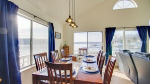 A dining area with a wooden table set for six, flanked by large windows with curtains, a view of the water, and a high ceiling with pendant lighting. - Lake View Cottage: Lake Ozarks Getaway (Gravois Mills)
