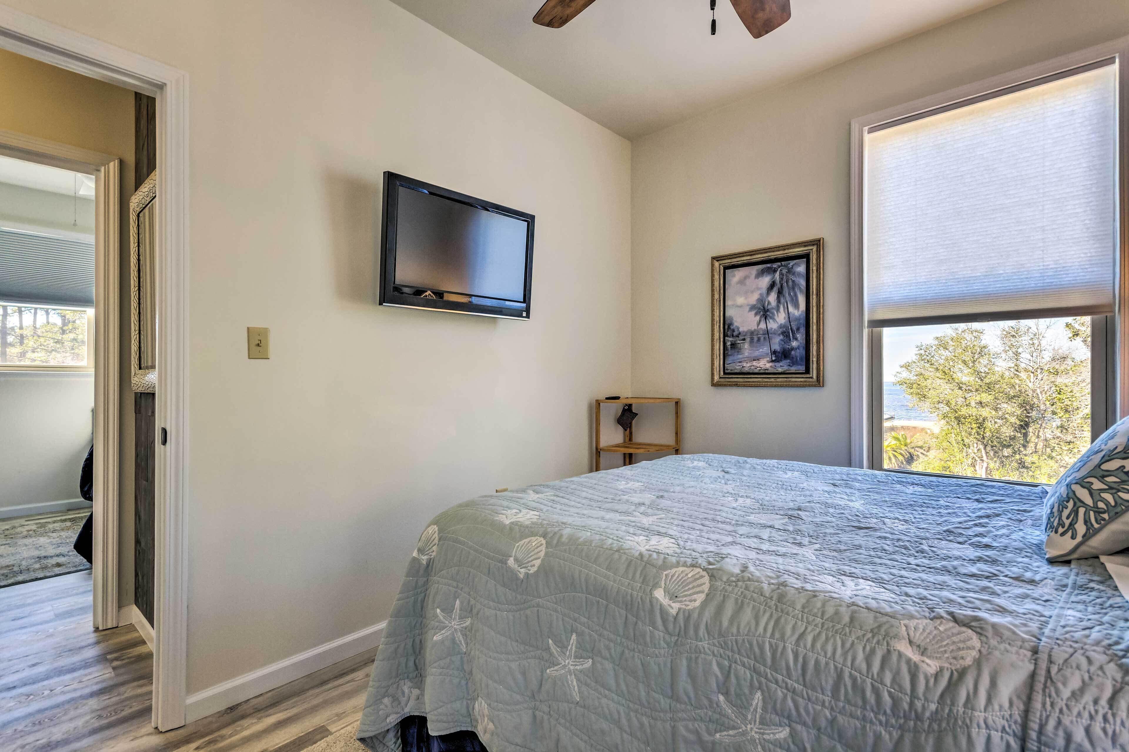 A bedroom with a bed, a TV mounted on the wall, a window with blinds, and a door leading to another room.
