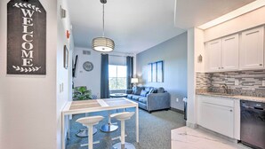 A modern kitchen with white cabinets and a built-in dishwasher, a dining area with a white table and bar stools, and a living room with a gray sofa and a blue couch. - Orlando Family Condo ~ 9 Mi to Magic Kingdom! (Orlando)