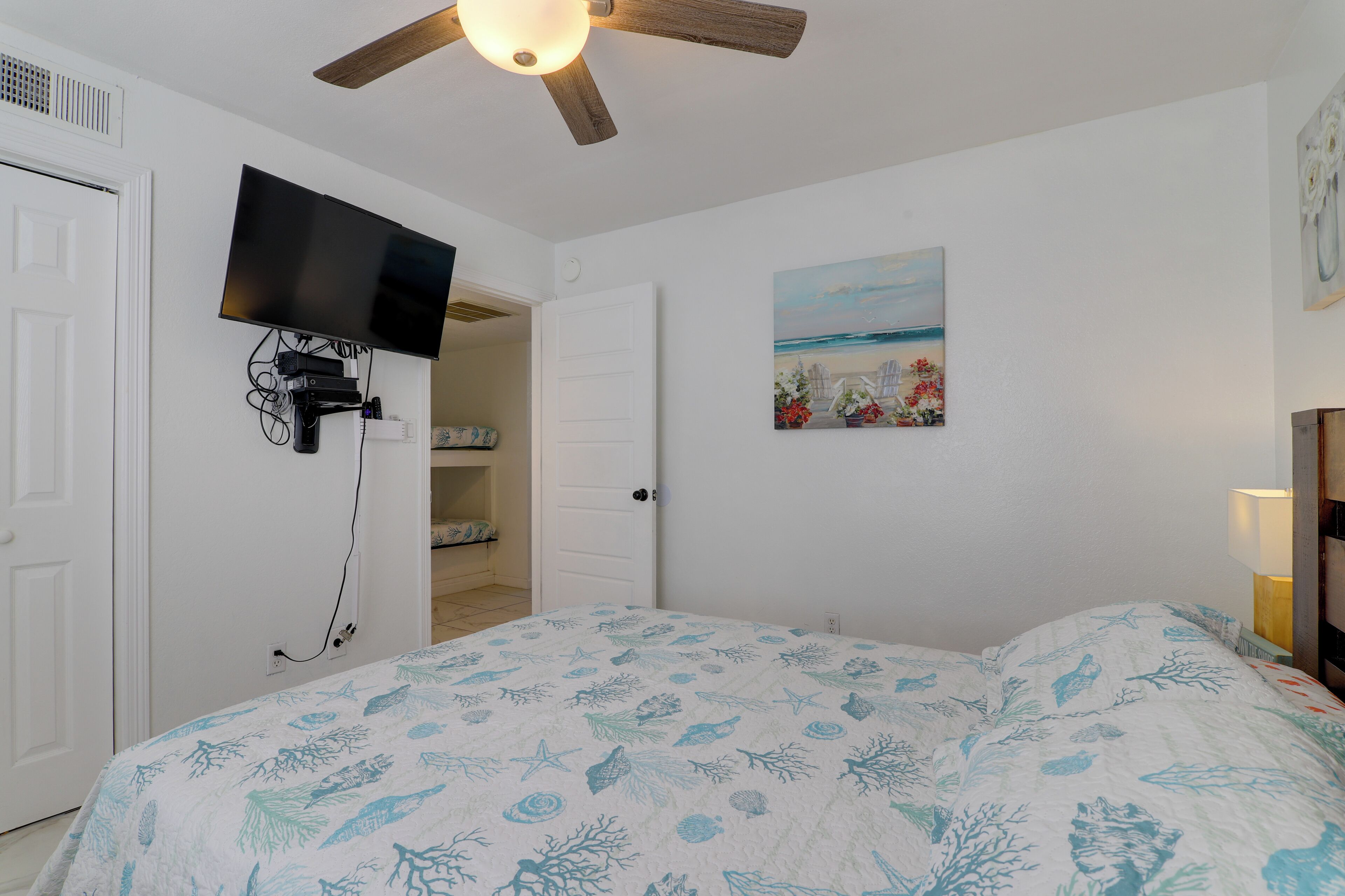 Un dormitorio con una cama, ventilador de techo, televisor montado en la pared y un cuadro.