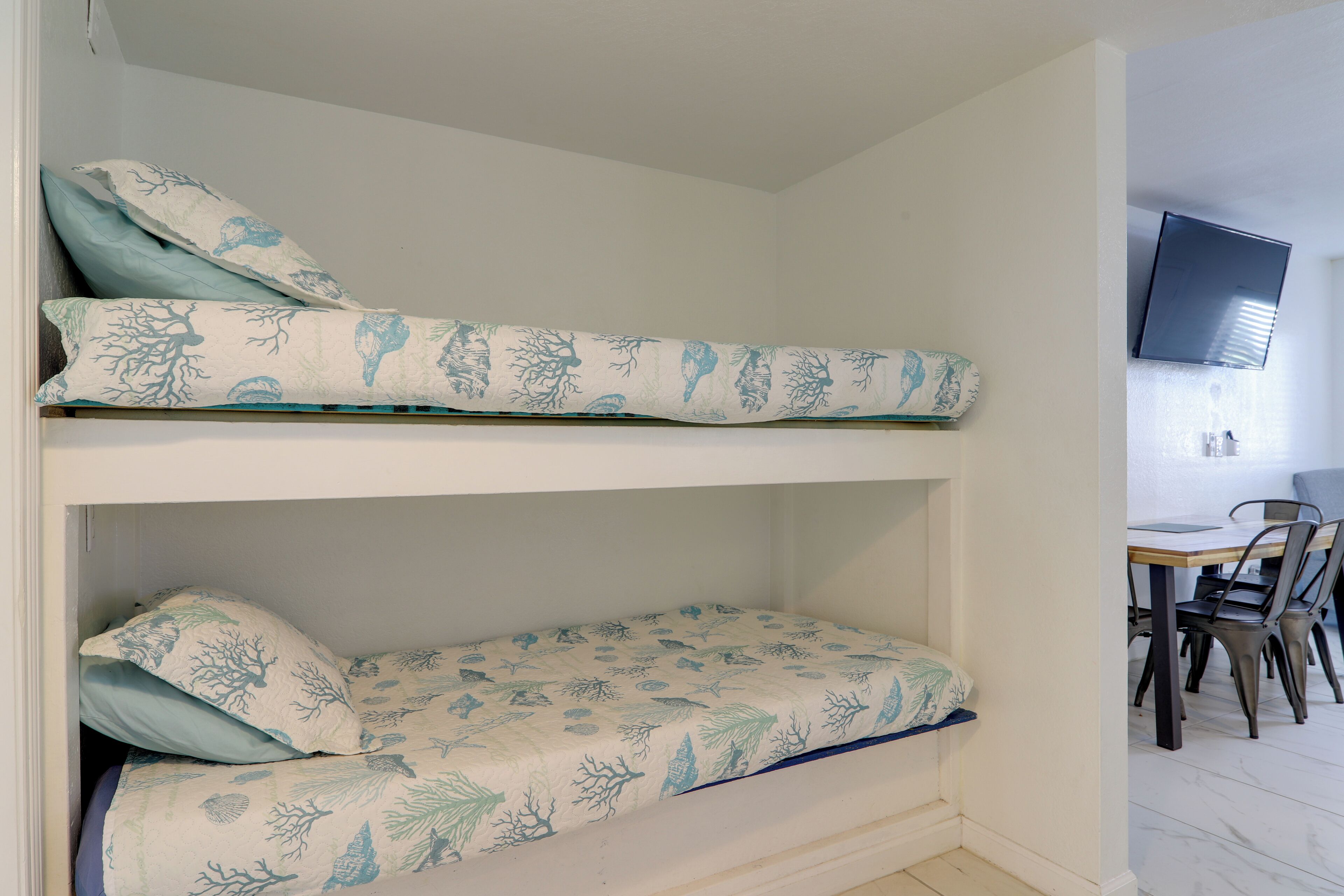 A bunk bed with patterned bedding, a television mounted on the wall, and a dining area with chairs and a table.