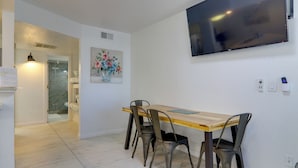 A dining area with a wooden table and metal chairs, a flat-screen TV mounted on the wall, and a painting of flowers. - Beach Digs: Walk to Restaurants, Bars & Ocean (South Padre Island)