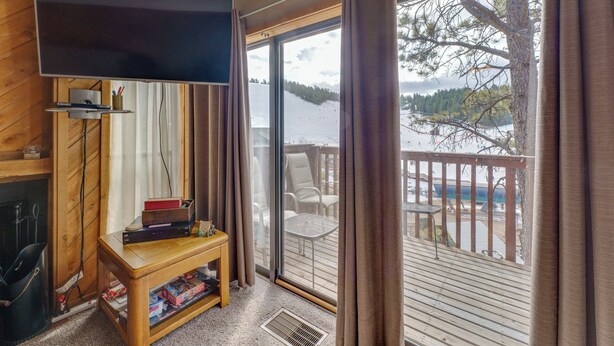 Et værelse med et fladskærms-tv, et træbord med bøger, en lille reol og en skydedør med glas, der fører ud til en balkon med havemøbler.