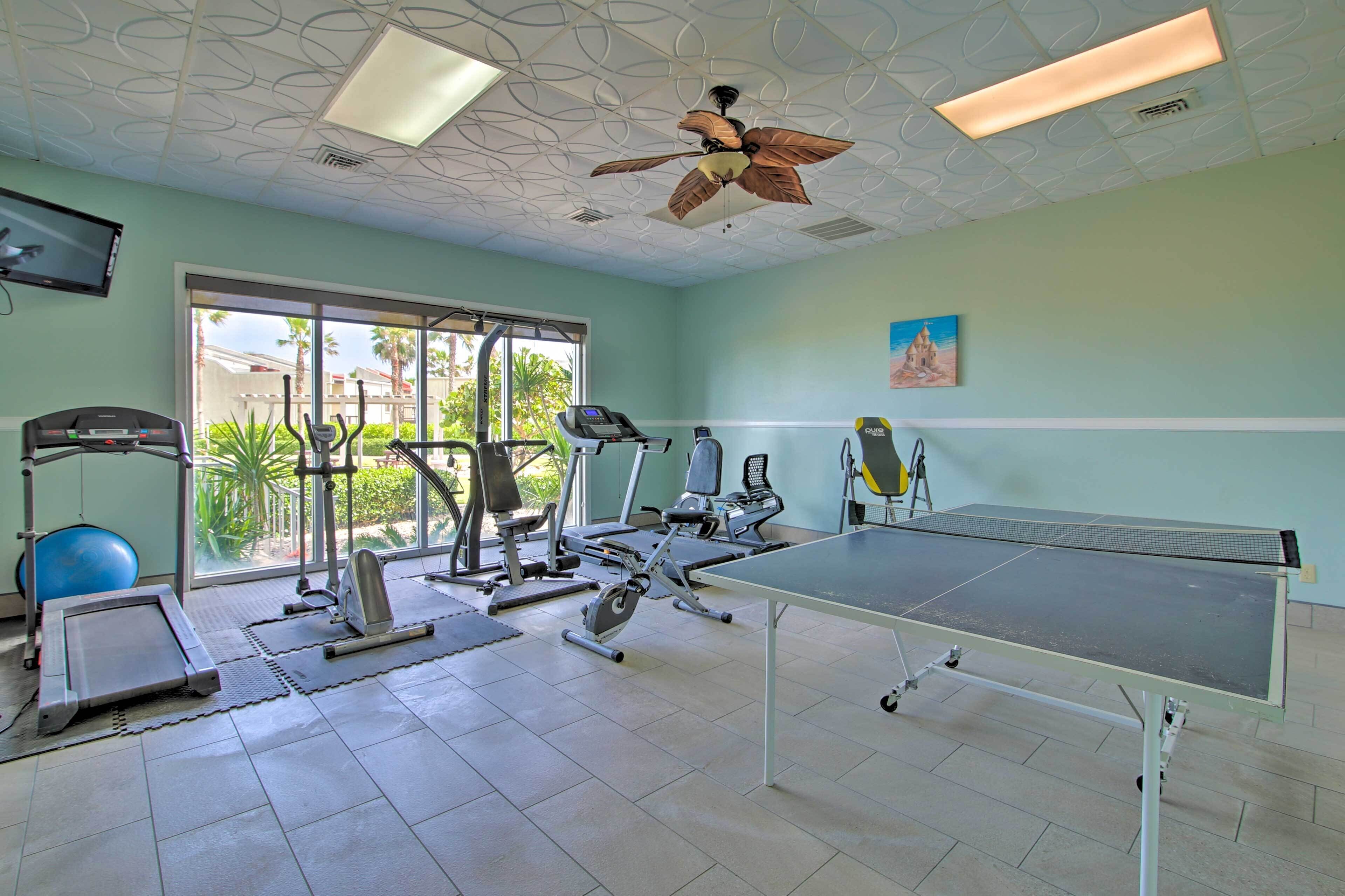 A gym with exercise equipment, a ping pong table, and large windows with a view of palm trees.