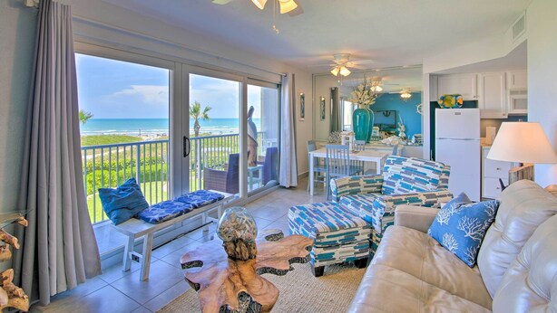A spacious living area with a balcony overlooking a beach, furnished with a sofa, chairs, and a dining table.