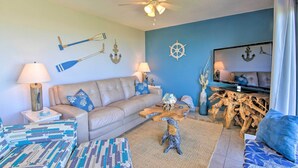 A living room with a beige sofa, two side tables with lamps, a wooden coffee table, and a television on a wooden stand. Nautical decor is visible on the walls.
