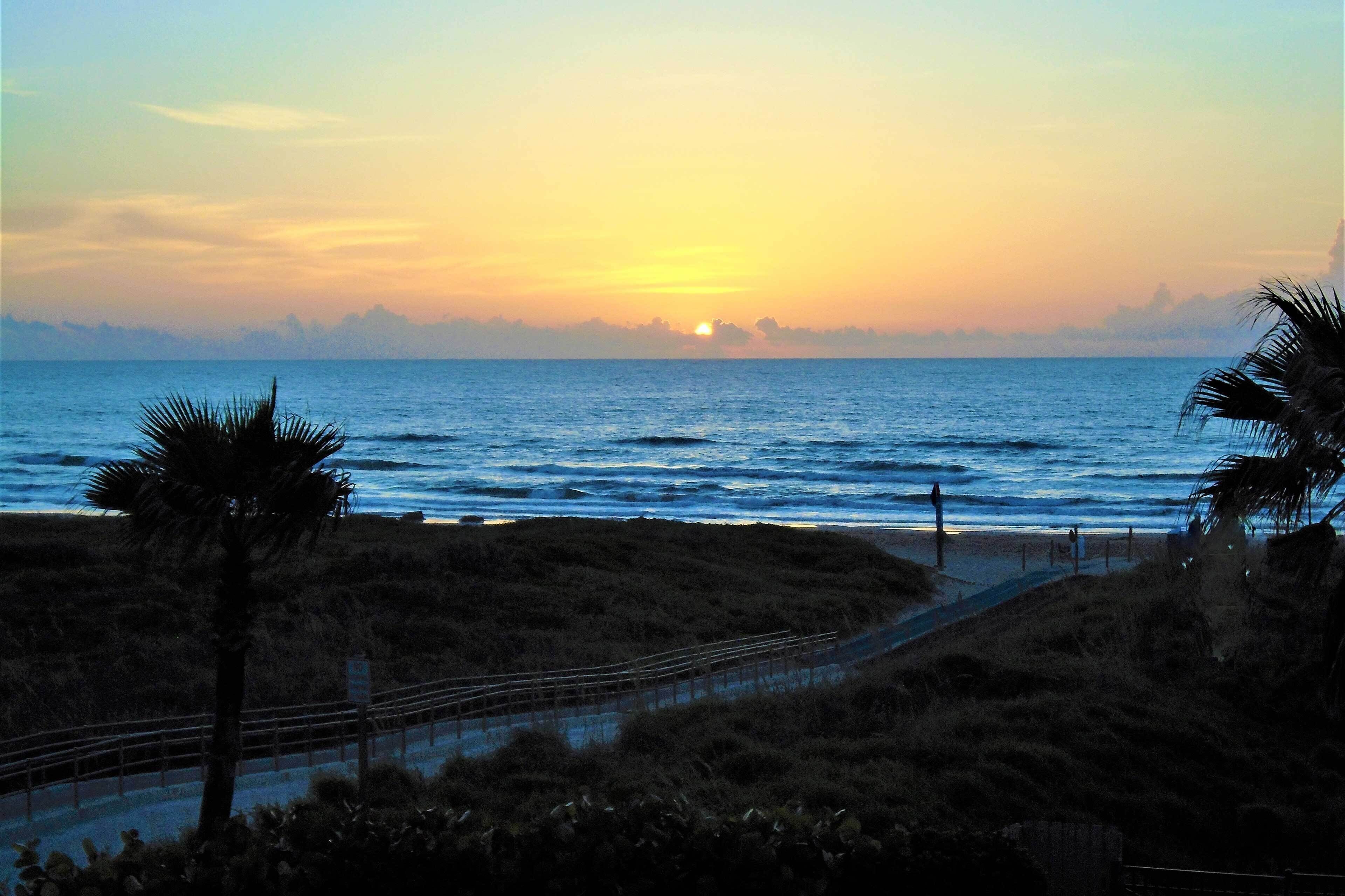 A sunset view over a beach with a pathway leading to the water, surrounded by vegetation.