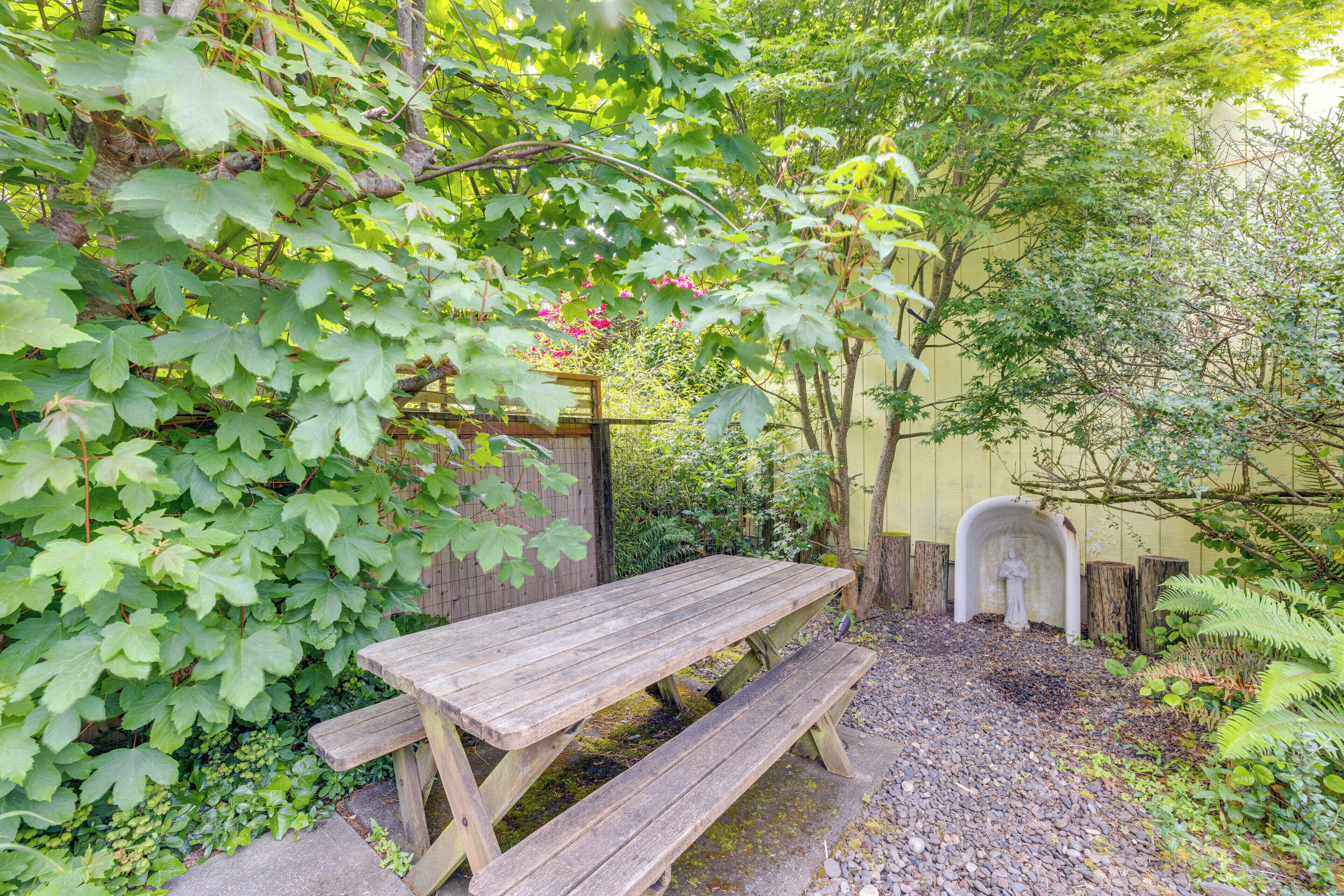Une table de pique-nique en bois, avec des bancs, se trouve dans un jardin où la végétation est luxuriante, et une petite statue est visible au fond.
