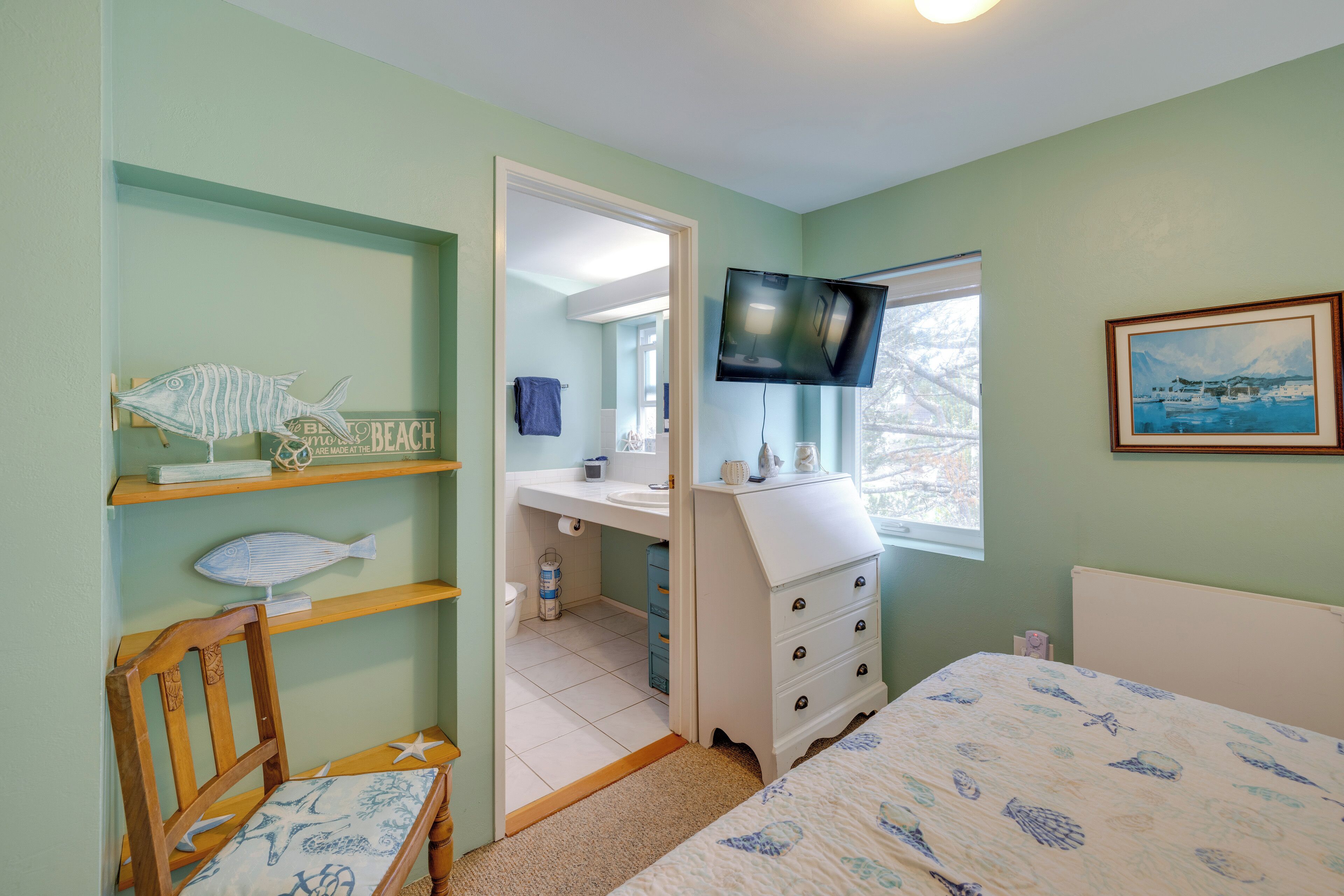 A bedroom with a bed, a wooden chair, a dresser, a television, and a window with a painting.
