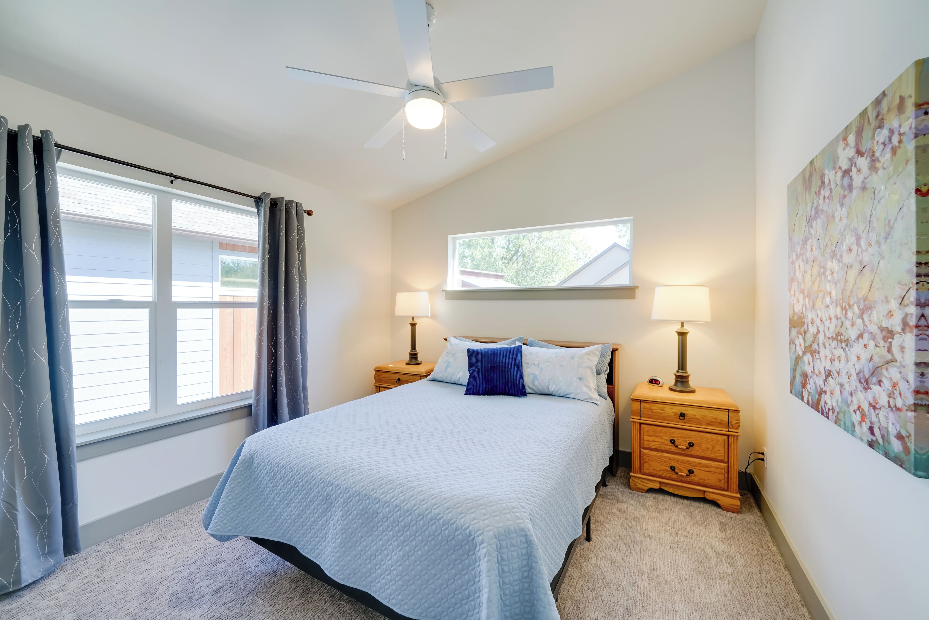 A bedroom with a bed, two bedside tables with lamps, a ceiling fan, a window with curtains, and a wall-mounted picture.