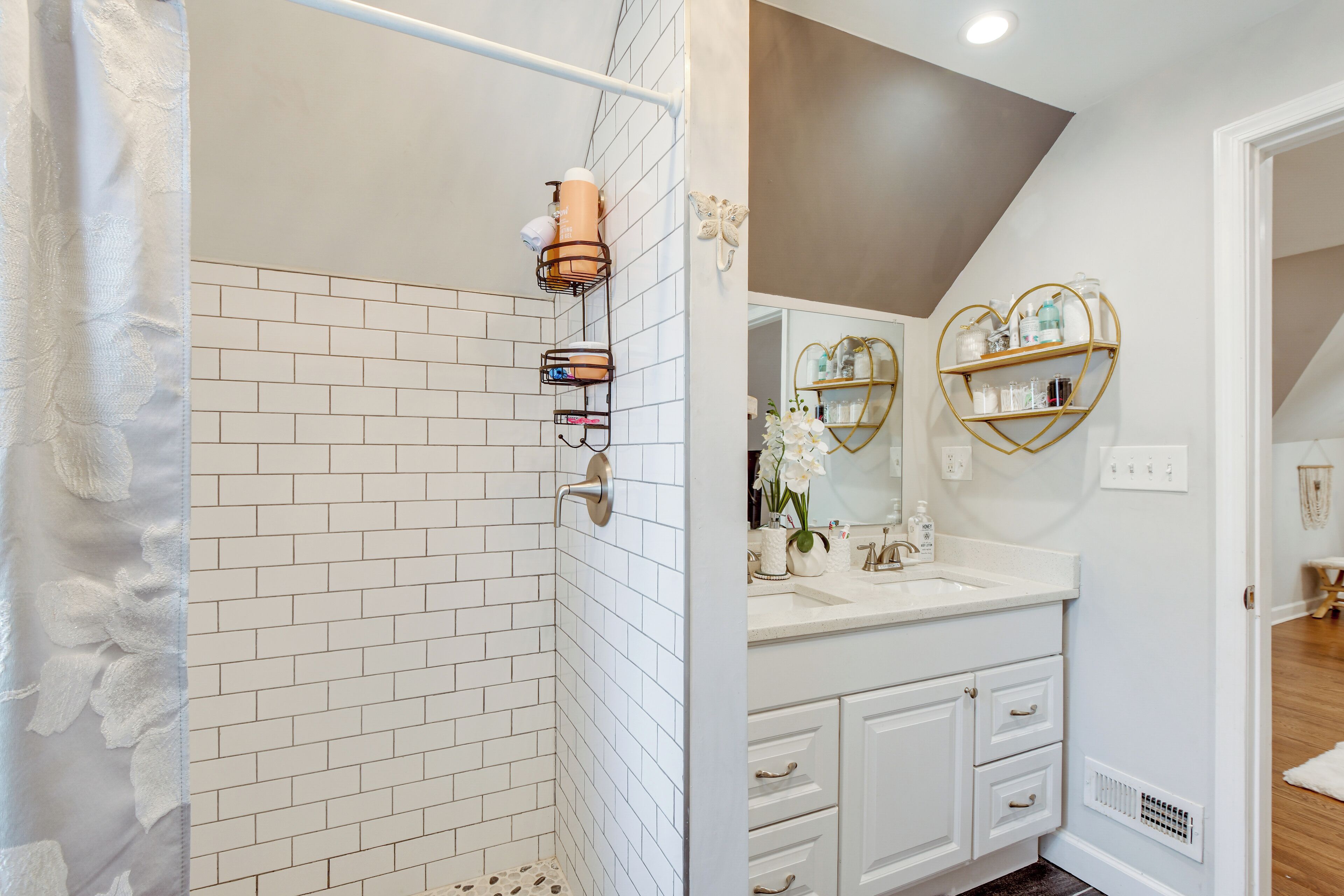 Une salle de bain avec des murs carrelés de blanc, un espace douche, un meuble-lavabo avec un miroir et une étagère en forme de cœur où sont rangés les articles de toilette.