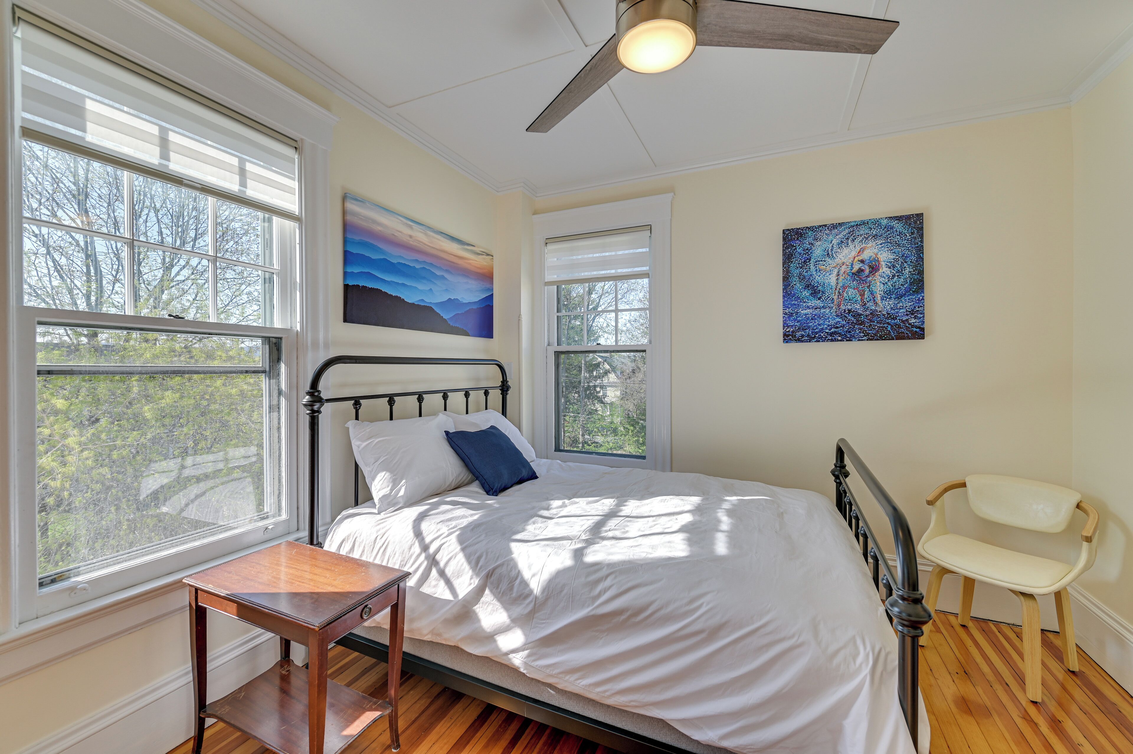 A bedroom with a bed, two windows, a ceiling fan, and two paintings on the walls.