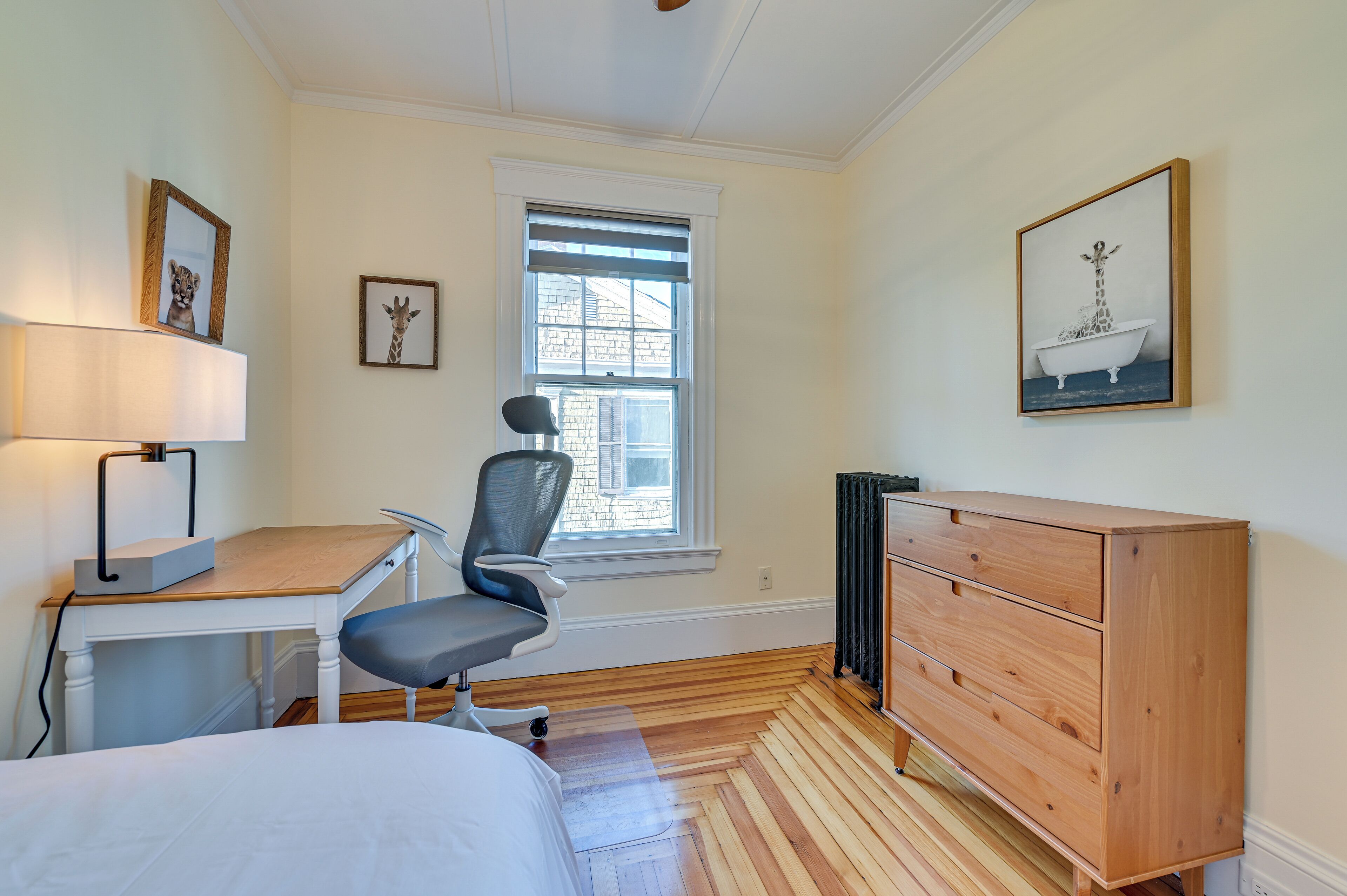 A bedroom with a bed, a desk with a lamp, a chair, a dresser, and a window with blinds.