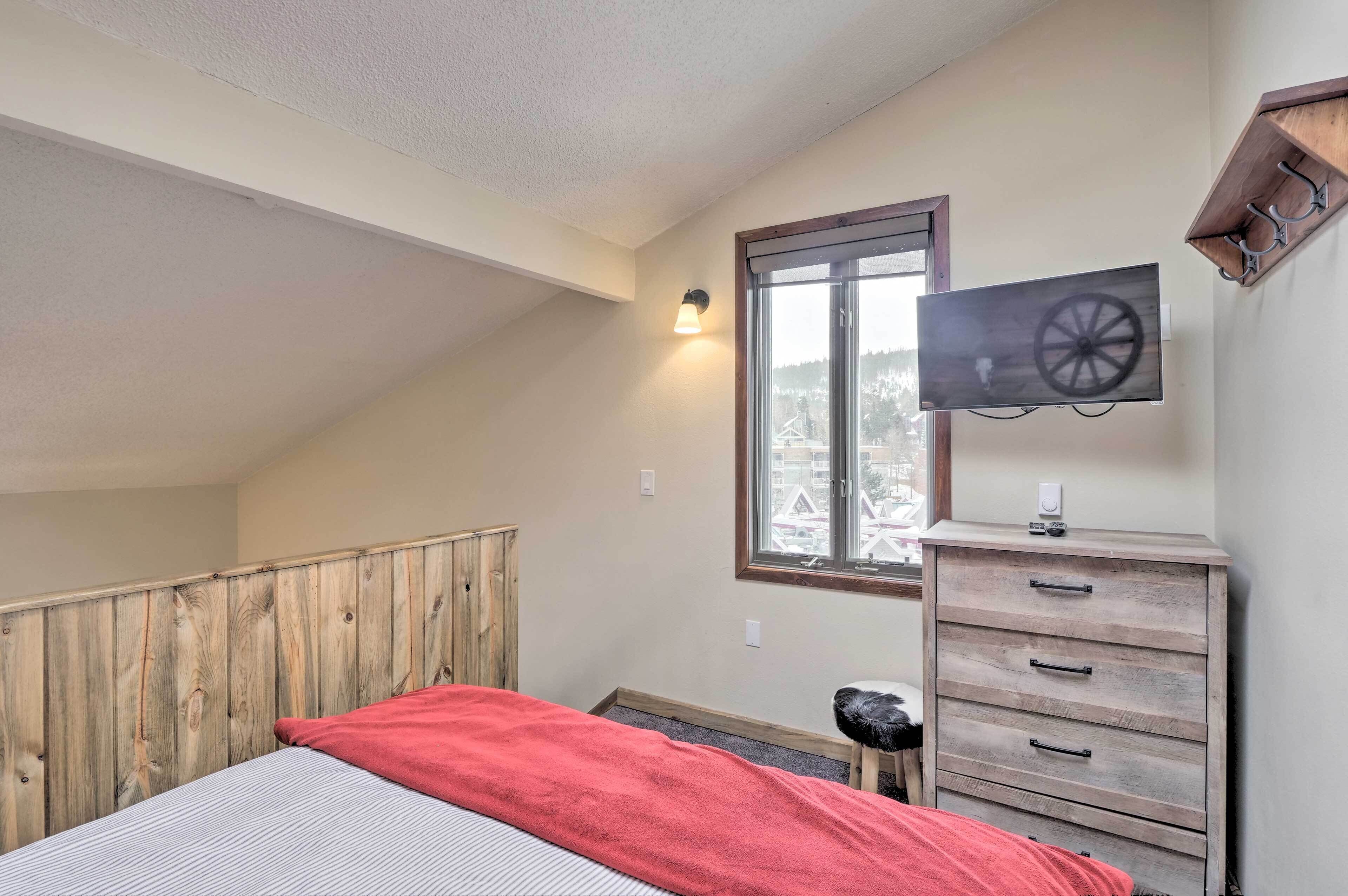 Une chambre avec un plafond en pente, un lit recouvert d’une couverture rouge, une commode en bois, un téléviseur fixé au mur et une fenêtre donnant sur des arbres.