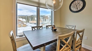 A dining area with a wooden table and chairs, a wall clock, and a view of a snowy landscape through large windows.