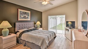 A bedroom with a bed, two bedside tables with lamps, a ceiling fan, a TV, and a large window with blinds. - 7 Mi to Wdw: Indian Ridge Home + Community Perks! (Kissimmee)