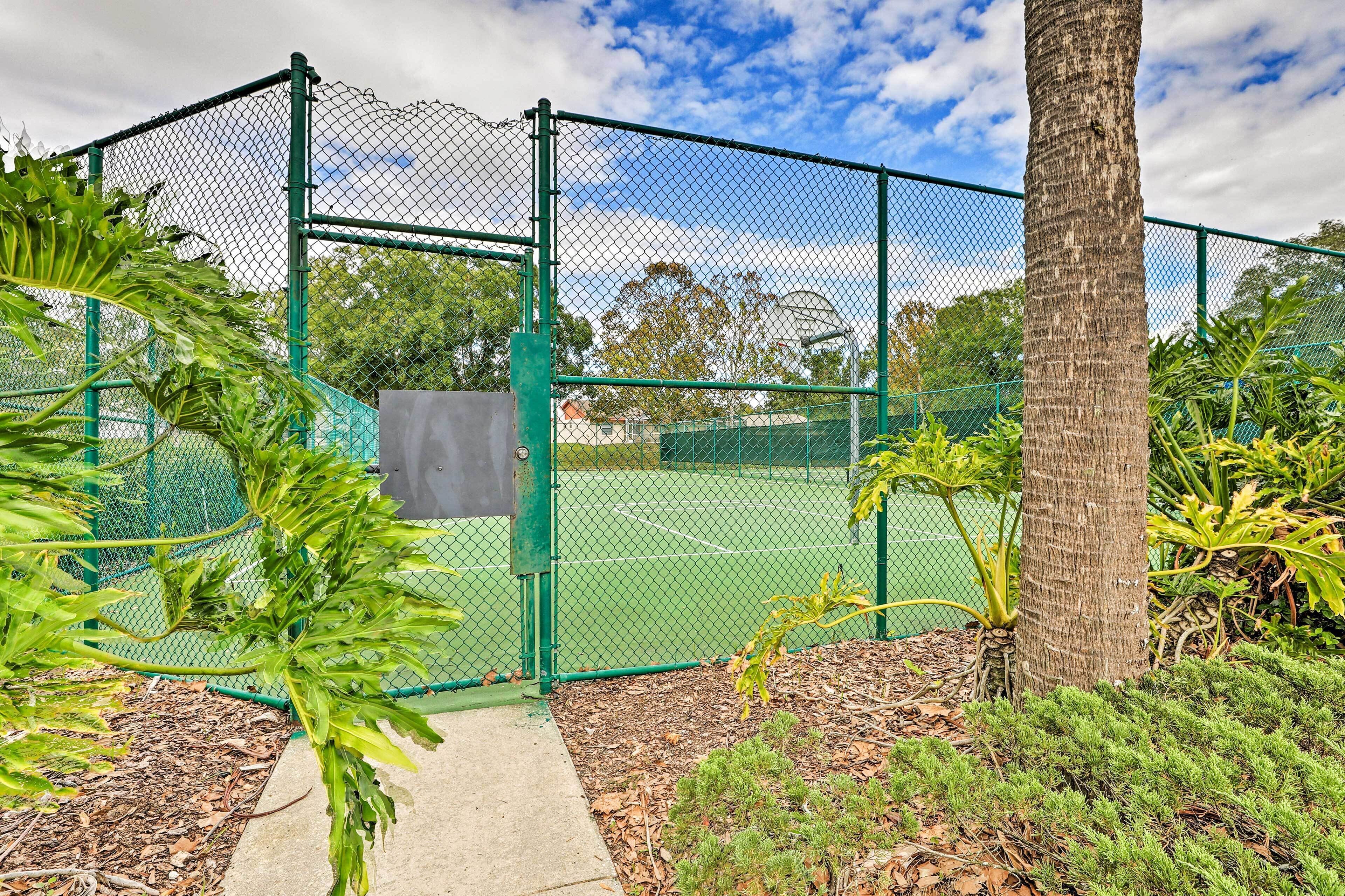 A tennis court enclosed by a green chain-link fence with a concrete pathway leading to the entrance.
