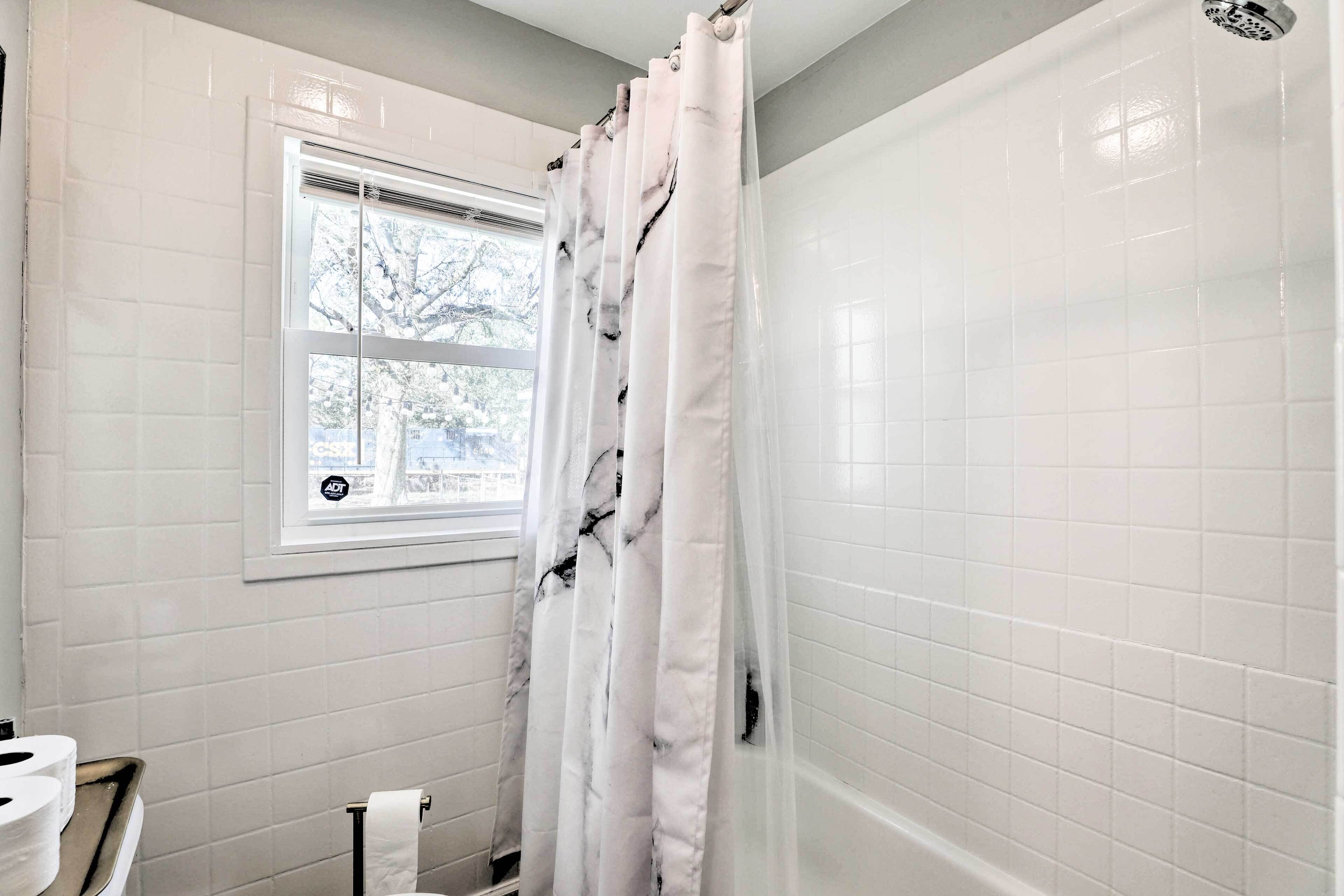 A bathroom with white tiled walls, a window with a view of trees, a shower curtain, and a towel rack.