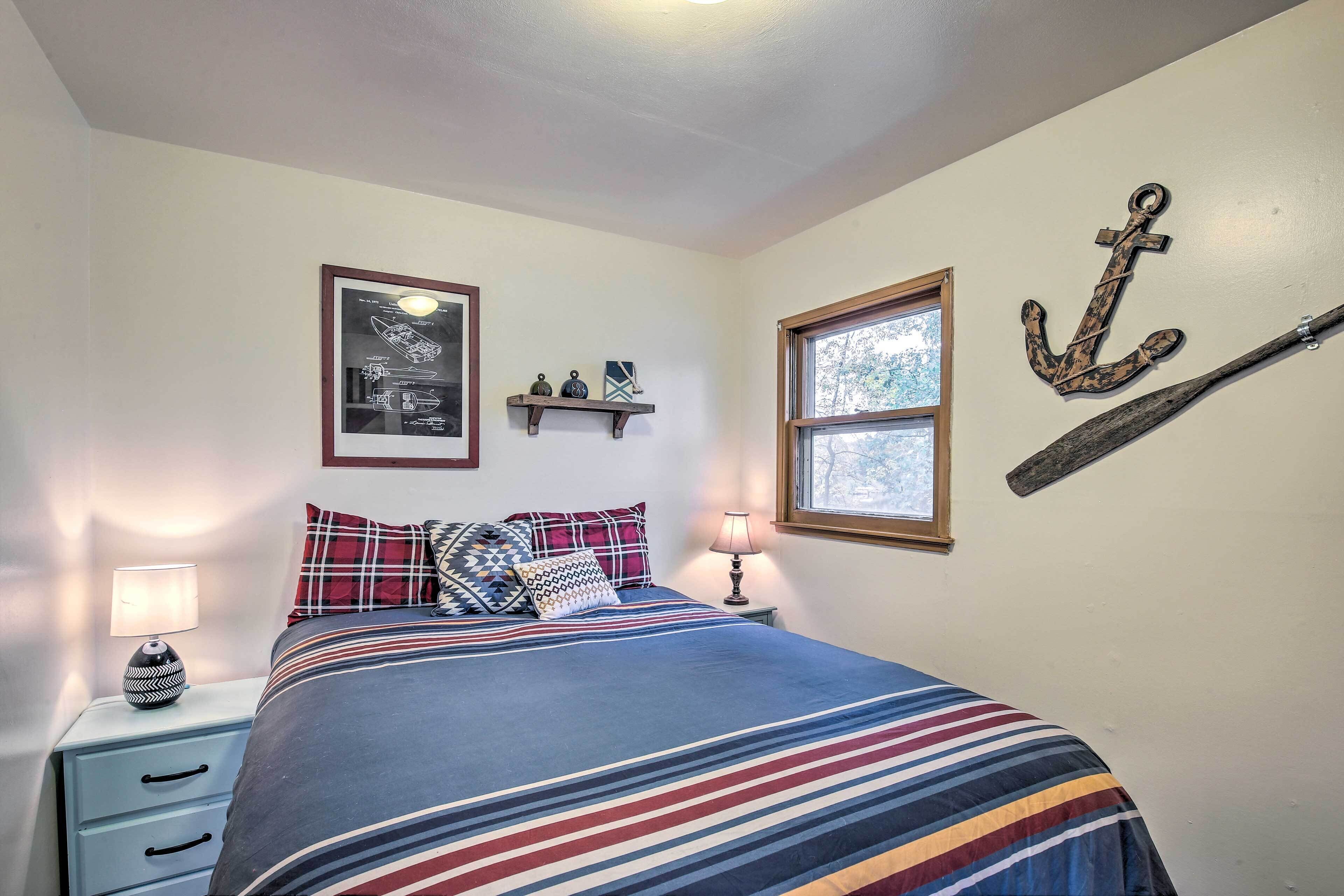 A bedroom with a bed, a nightstand, a lamp, a framed picture, a window, and a wall-mounted anchor.