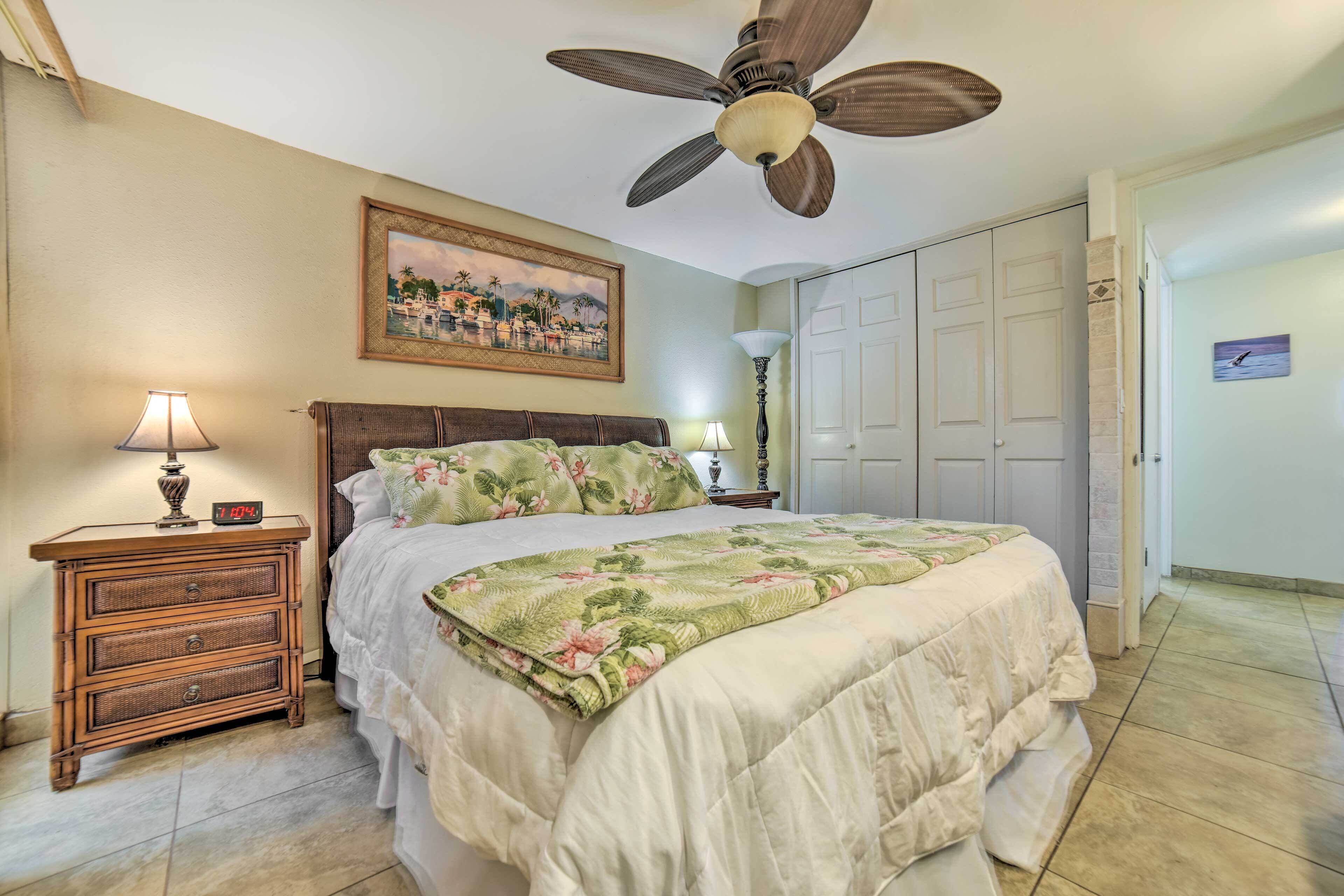 A bedroom with a bed, a nightstand with a lamp, a ceiling fan, a painting on the wall, and a door.