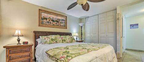 A bedroom with a bed, a nightstand with a lamp, a ceiling fan, a painting on the wall, and a door.