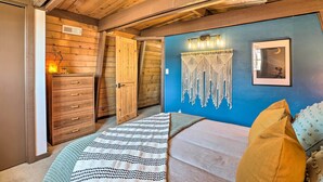 Ein Schlafzimmer mit Holzboden, einer blauen Akzentwand, einem Bett mit gemustertem Überwurf, einem Wandbehang aus Makramee und einem gerahmten Bild.