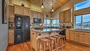 A spacious kitchen with wooden cabinets, a large island with stools, a refrigerator, and a view of mountains through large windows. - Mountain-view Deck: South Lake Tahoe Retreat! (South Lake Tahoe)