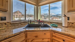 A kitchen with a granite countertop, wooden cabinets, a stainless steel sink, and a view of snow-capped mountains through large windows. - Mountain-view Deck: South Lake Tahoe Retreat! (South Lake Tahoe)