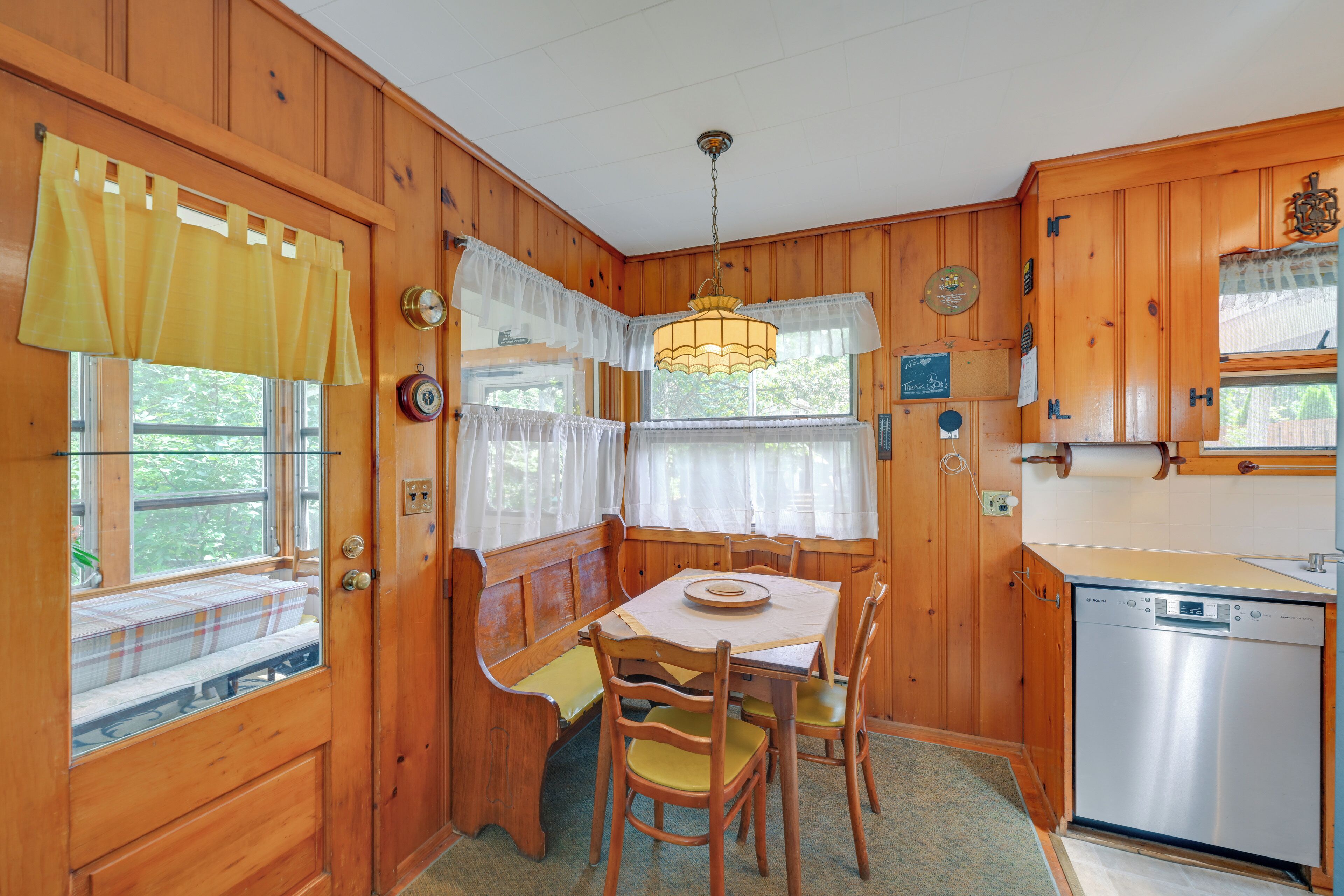 A wooden cabin interior with a dining table, chairs, a bench, and a kitchen area with a dishwasher.