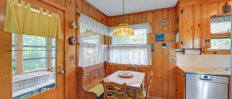 A wooden cabin interior with a dining table, chairs, a bench, and a kitchen area with a dishwasher.