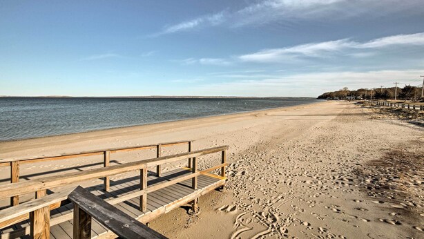 A wooden pier leading to a sandy beach with footprints, a clear sky, and a calm sea. - Walk to Beach: Long Island Wine Country Home! (Cutchogue)