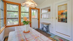 A room with a plaid tablecloth, a white vase with flowers, a vintage sewing machine, and a door leading to a kitchen. - Walk to Beach: Long Island Wine Country Home! (Cutchogue)