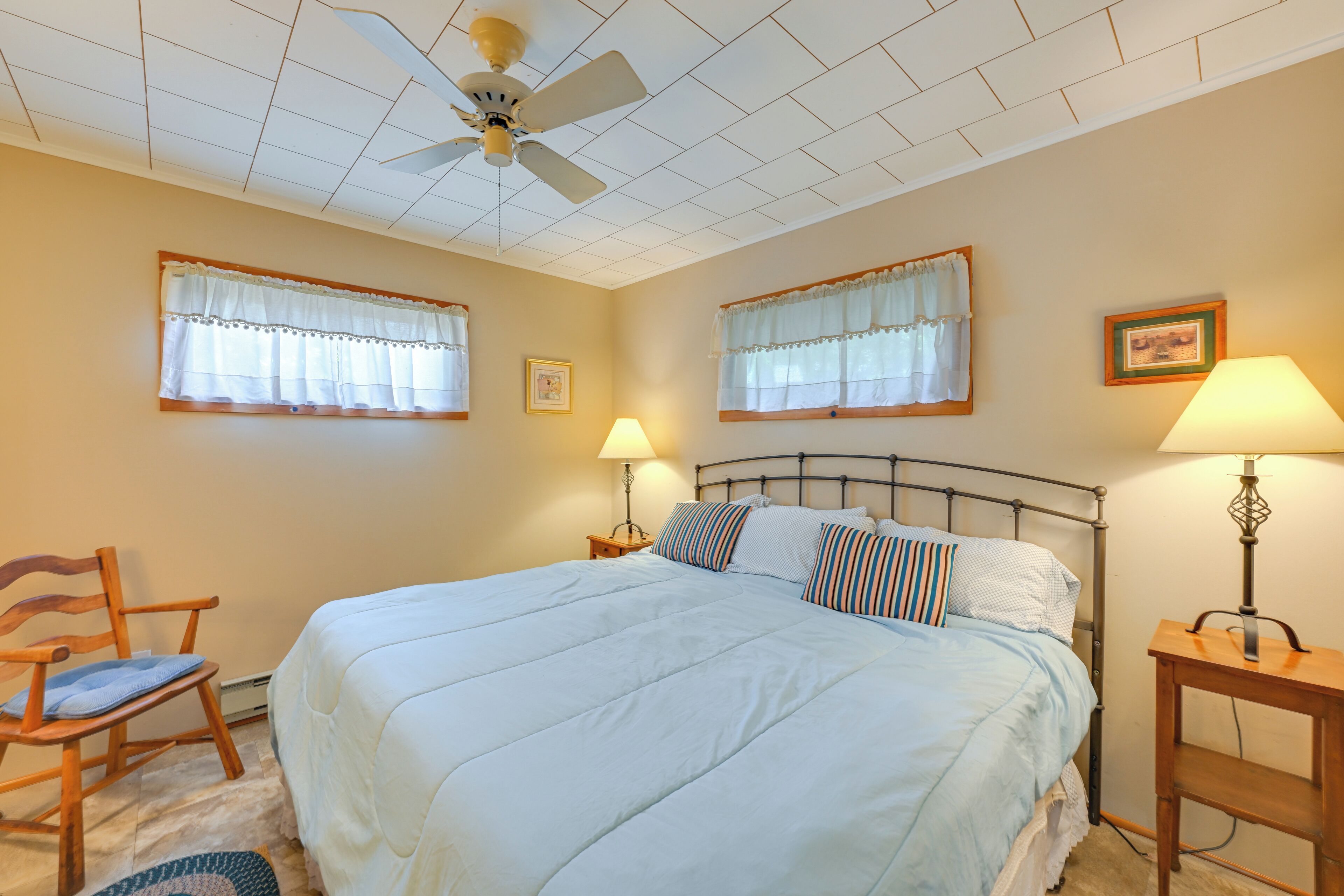 A bedroom with a bed, two bedside tables, a chair, a ceiling fan, and two windows with curtains.