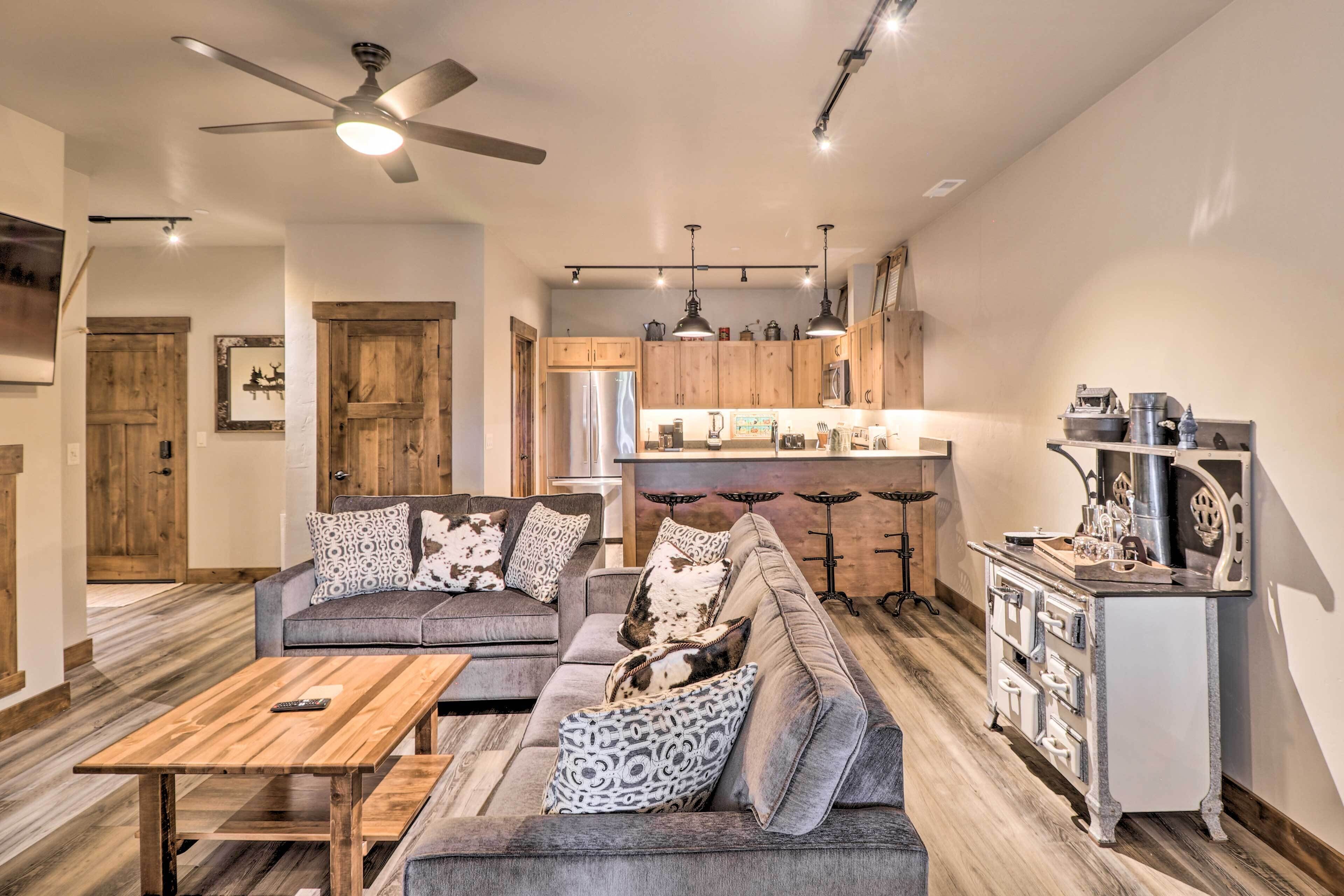 A modern living room with a sectional sofa, wooden coffee table, and a kitchen area with wooden cabinets and a refrigerator.