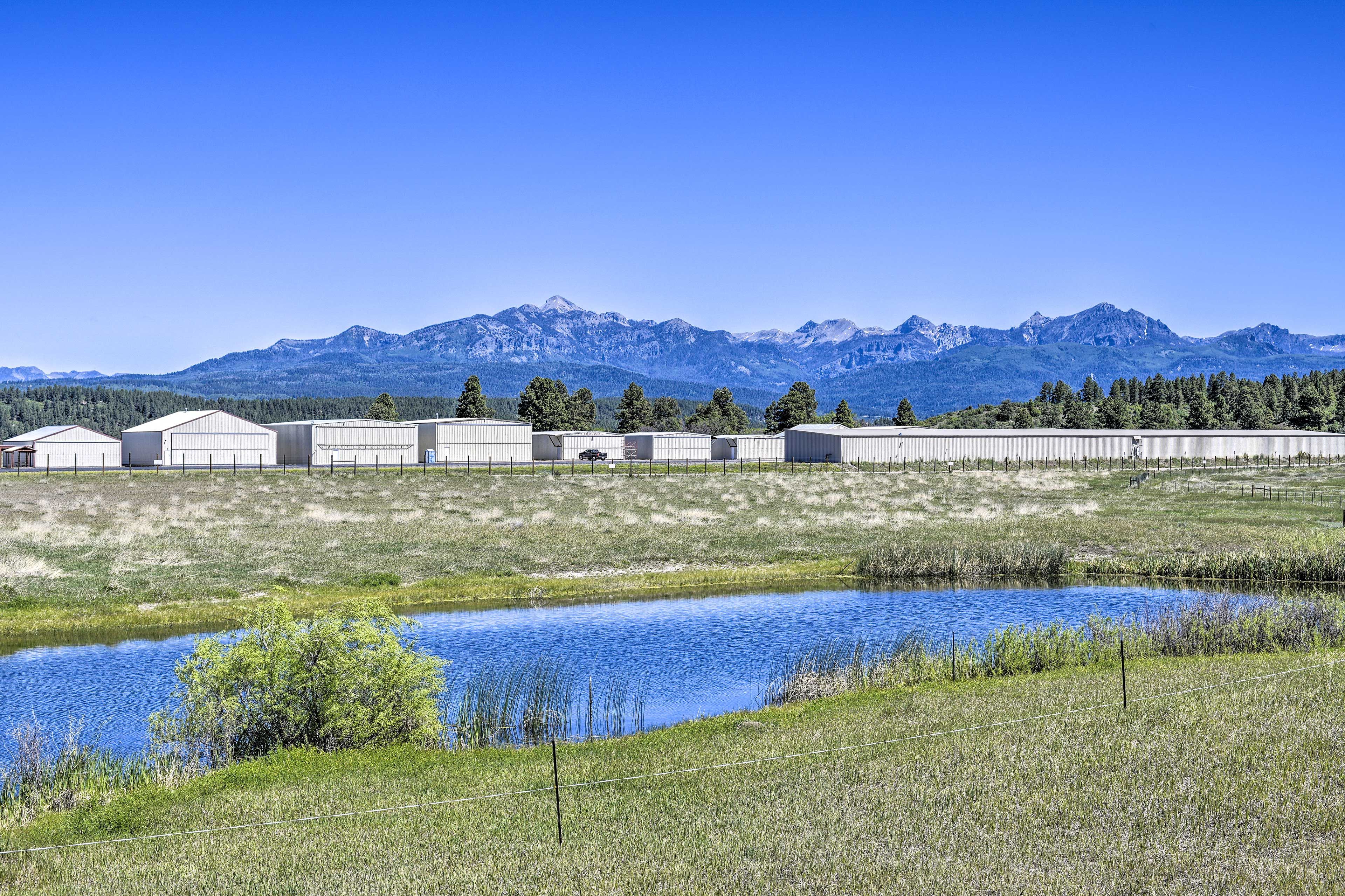A rural setting with a row of buildings, a small pond, and mountains in the background.
