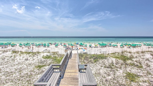 Ein Strand mit einer Holzpromenade, die zum Ufer führt, umgeben von Strandsonnenschirmen und Menschen.
