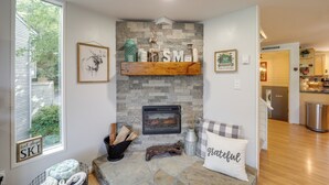 A cozy living room with a stone fireplace, a wooden shelf with decorative items, and a view of the outdoors through a large window. - Walk to Ski & Golf: Sugar Mountain Condo! (Banner Elk)