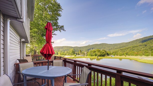 A deck with a table and chairs, overlooking a lake and hills. - Walk to Ski & Golf: Sugar Mountain Condo! (Banner Elk)