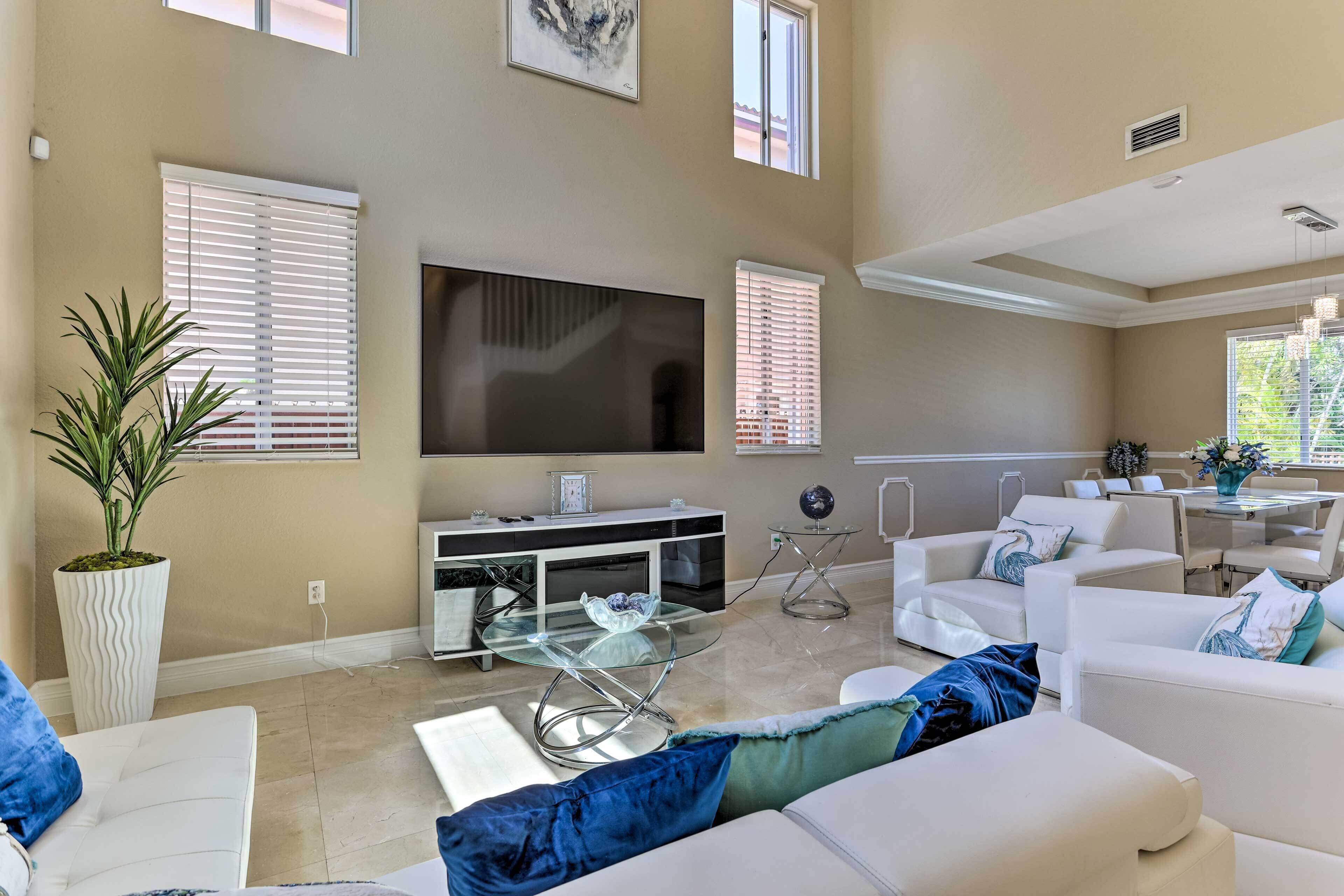 A spacious living room with a large flat-screen TV, white sofas, a glass coffee table, and a potted plant.