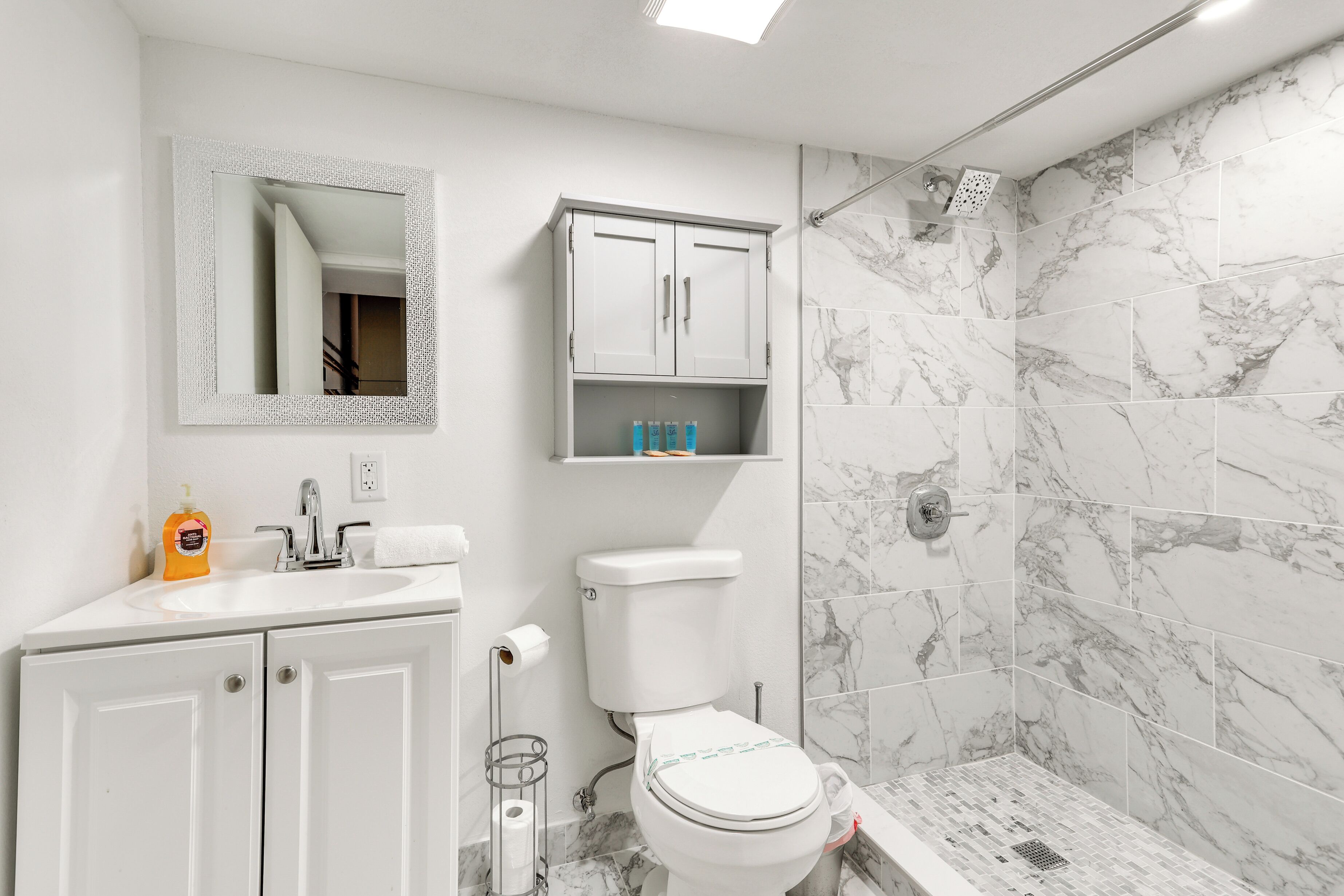 A bathroom with a toilet, sink, mirror, cabinet, and shower area with marble tiles.