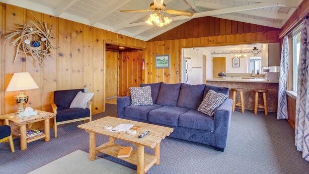 L’intérieur d’un chalet accueillant, avec un plafond en bois, un canapé bleu, une table basse en bois et un coin cuisine visible à travers une porte ouverte.